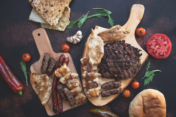 Various grilled meats served on wooden cutting boards with fresh vegetables and bread on a dark...