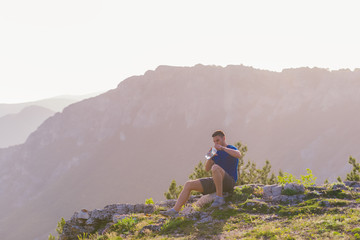 Fototapeta premium Handsome athlete sitting at a rocky peak while looking at the breathtaking mountain line and a beautiful lake while wearing a blue shirt and grey shorts.