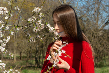 a girl in red golf sniffs and enjoys the aroma of cherry blossoms. a white face mask on the chin to protect against coronavirus, viruses and diseases. Protection and personal hygiene on the street.