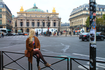 Obraz premium Blonde girl in a brown coat in front of the Palace or Opera Garnier and the National Academy of Music. Automobiles and pedestrians in front of the Grand Opera.