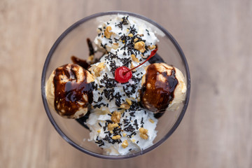 Sweet dessert seen from above served in a glass consisting of vanilla ice cream, brownie pieces, cream, chocolate syrup and chocolate shavings with a cherry on top.