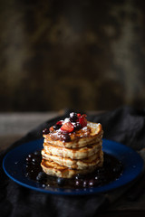 Thick pancakes are stacked on a plate covered in blueberries, strawberries and syrup ready for breakfast or brunch