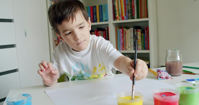School boy reaching for paints with paintbrush in his hand and painting. Child with paintbrush and colorful paints at home. Hand-held shot, single shot, medium close up, 4K.