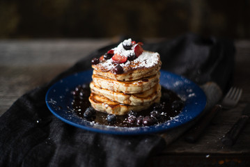 Thick pancakes are stacked on a plate covered in blueberries, strawberries and syrup ready for breakfast or brunch
