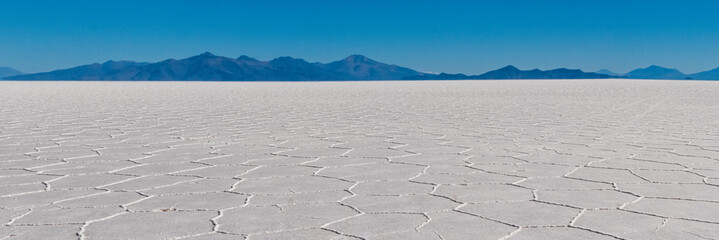 Panoramic view of the Uyuni salt dessert with hexagonal salt formations and mountains at the Salar de Uyuni (Uyuni Salt Flat) during daytime, Bolivia. - obrazy, fototapety, plakaty