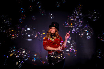 Girl in stage costume and top hat on her head. Female magician, an illusionist in theatrical clothes, makes show with soap bubbles on black background. Concept of theatrical performance