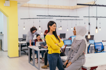 Portrait of two African American businesswomen talking to each other while standing in a modern business office with their colleagues, coworkers in the background. Multi-ethnic society..
