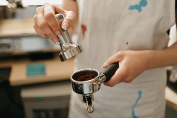 A female barista holds a metal tamper and a portafilter with coffee in a coffee shop. A barista preparing for pressing ground coffee for brewing espresso or americano in a cafe.