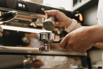 A close photo of female hands holding a metal tamper and a portafilter with coffee in a coffee shop. A barista preparing for pressing ground coffee for brewing espresso or americano in a cafe.