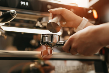 A close photo of female hands which holds a metal tamper and a portafilter with coffee in a coffee shop. A barista preparing for pressing ground coffee for brewing espresso or americano in a cafe.