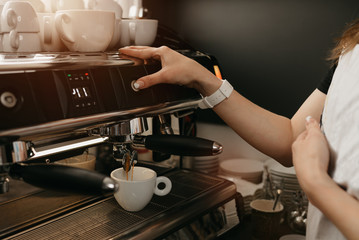 A female barista with white apron brewing espresso in a professional espresso machine in a coffee shop. A close-up photo of pouring a coffee in a white cup from a coffee machine in a cafe.