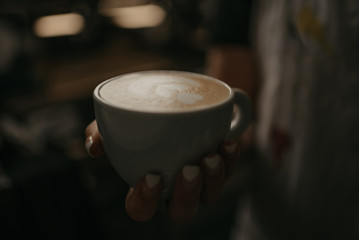 A female barista holds a cup of hot latte in her hand in a cafe. A waitress preparing a customer order in the evening.