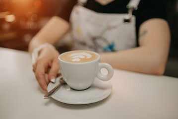 A female barista holds out a cup of fresh latte with a saucer and spoon to a client in a cafe. A barista preparing a customer order.