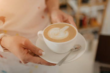 A close-up photo of a cup of latte which a female barista holds in her hands in a coffee shop. A barista preparing an order in a cafe.