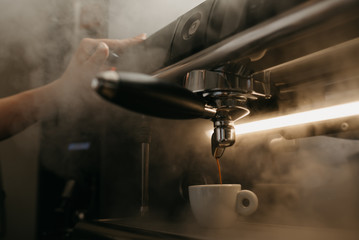 A close photo of a professional coffee machine. A barista pours an espresso from a coffee machine in a white cup. Steam around.