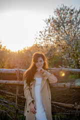 A young woman in a white dress and beige trench coat stands near a wooden fence in nature, a beautiful girl with long hair in the countryside