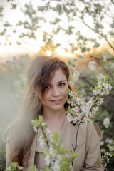 Fototapeta premium A young girl with long hair in a beige trench coat and white dress next to a flowering tree, girl's face in flowers