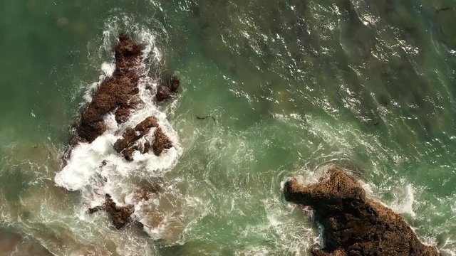 Aerial top view of waves break on rocks in a blue ocean. Sea waves on beautiful beach aerial view drone 4k shot. Bird's eye view of ocean waves crashing against an empty stone rock cliff from above.