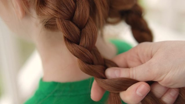 Caring mommy braiding pigtail to her little red-haired daughter, love and care