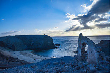 Fototapeta na wymiar Papagayos beach in Lanzarote at sunset with sun bursting from clouds, arched rock formation and silhouetted people on beach and cliffs