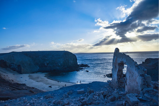 Papagayos beach in Lanzarote at sunset with sun bursting from clouds, arched rock formation and silhouetted people on beach and cliffs