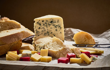 Piece of cheese with blue mold and other kinds of sliced small cubes on parchment with fresh baguette and sausage, wine glasses. Gourmet still life.