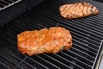Raw steak slathered in sauce alongside cooked steak grilling on barbecue