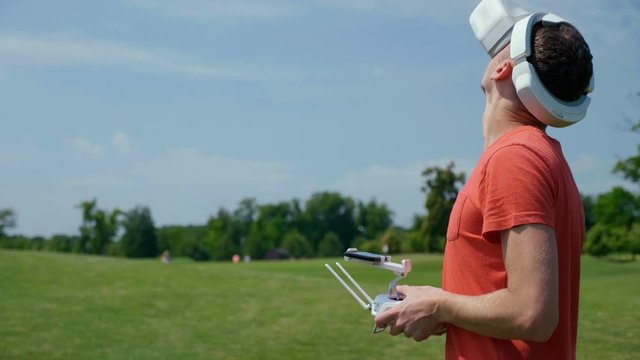 Man controls a quadrocopter through a remote control and looks at the video with goggles on his head. Drone operator in a red T-shirt and blue shorts in park is filming drone video