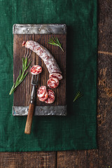 Dried sausage on a rustic board with rosemary and spices on a green tablecloth. A piece of sausage is pinned on the fork. German meat products. View from above. Rustic food.