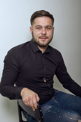 Fototapeta premium Young Bearded Dark Haired Man In Black Stylish Shirt On Gray Background, Confident Serious Man Sitting On Chair And Looking Straight. Concept Of Ordinary Ordinary People Who Work In An Office