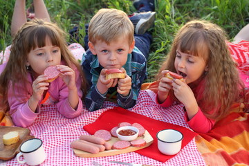 Three children at a picnic eating sausage sandwiches