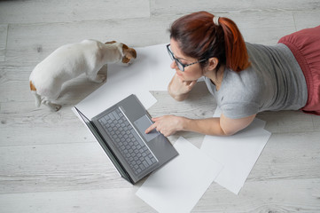 A woman lies on her stomach on the floor and is typing on a laptop. Girl freelancer works remotely from home with a puppy. Jack Russell Terrier next to the owner in a comfortable position.