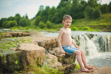 A boy in jeans shorts sits on a stone near a waterfall