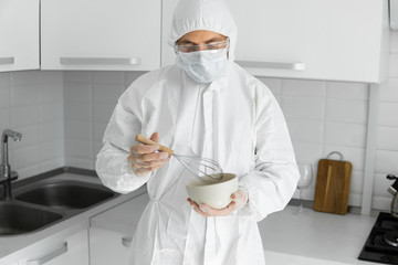 Man in white protective suit and in face medical mask cooks in the white kitchen at home during coronavirus. Stay at home. Enjoy cooking at home. COVID-19 global pandemic.