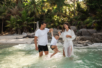 Happy smile european family have rest and run on white sand beach. Father, long black chestnut hair mother, blond baby boy. white cotton clothes. boho dress. t-shorts.Thailand. Aquamarine crystal sea