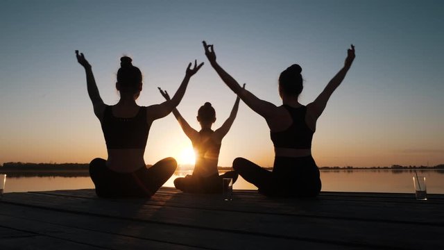 Silhouette of diverse group of three female doing yoga awe exercise sitting in lotus position together on boardwalk near calm water at dusks outdoor. Tranquil namaste for emotional relief, stress free