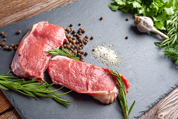 Raw beef meat and spices. Slices of fresh raw meat, rosemary and fresh herbs on a black slate stone plate. Preparing and marinating barbecue meat.