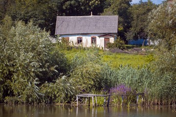 shabby and worn old country house ruining lonely, abandoned home and yard on bank of countryside lake, desolation and depression mood