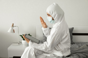 Man in protective white suit and medical mask is working from home on a bed with laptop and showing hello to his friends or business partners because of coronavirus epidemic.