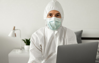 Man in protective white suit and medical mask is working from home on a bed with laptop because of coronavirus epidemic. Remote work during pandemic. Stay home during COVID-19 quarantine concept.