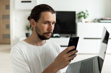 Man with beard, working at home with a laptop in front of him and speaking with a friends or business partners or clients with a smartphone and sitting on a floor. Freelancer working from home.