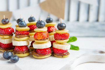 Mini pancakes canape with berries, chocolate cream and banana, new trendy social media food. Party healthy dessert bar on white table. Close up view, copy space. Small pancakes with fresh fruits.