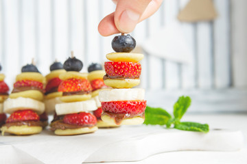 Mini pancakes canape with berries, chocolate cream and banana, new trendy social media food. Party healthy dessert bar on white table. Close up view, copy space. Small pancakes with fresh fruits.