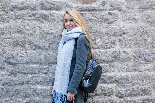 Portrait of young woman with light blue scarf and backpack standing in front of stone wall