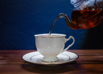 From a glass teapot, he pours black tea into a white porcelain cup. Wood surface and blue walls