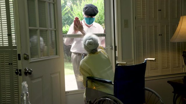 An elderly woman in a wheelchair social distancing because of COVID19 visits with a caring relative or neighbor through her glass storm door.