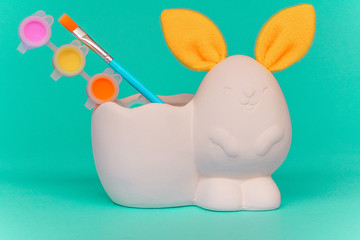 statuette of white figure of bunny or rabbit with yellow ears for decorating, Easter DIY with children. Multi-colored paints and brushes on green background. Minimal easter concept