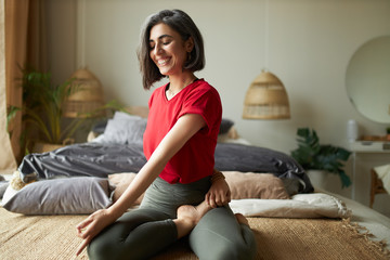 Cheerful young athletic woman sitting barefooted on carpet, keeping legs crossed, doing seated spinal twist during yoga class, enjoying stretching exercise, taking deep breaths, closing eyes