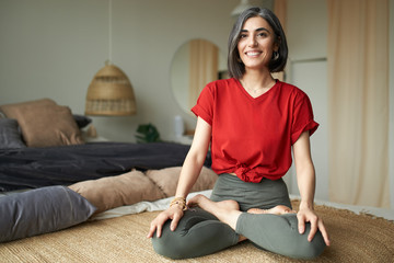 Premature graying of hair and body positivity concept. Attractive stylish young female sitting in cozy interior doing yoga, sitting in lotus posture, smiling broadly, enjoying complicated asana