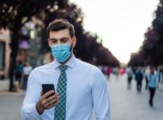 Young elegant businessman stands in middle of crowded street with surgical mask looking a phone